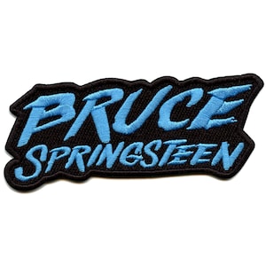 May include: A black embroidered patch with the name "BRUCE SPRINGSTEEN" in light blue, bold lettering. The patch has a black border and is designed to be sewn or ironed onto clothing or accessories.