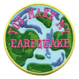 Op de afbeelding: Een ronde, geborduurde patch met een blauw en groen aardeontwerp. De tekst "You Make My Earfquake" is in roze rond de rand van de patch geschreven.