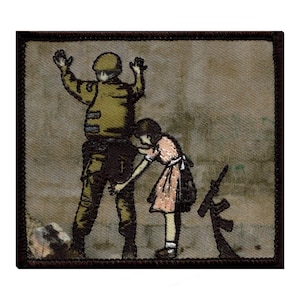 May include: A brown and tan embroidered patch depicting a soldier with their hands raised and a young girl reaching out to them. The soldier is wearing a green uniform and a helmet. The girl is wearing a pink dress. A black and white stylized rifle is in the background.