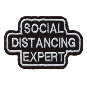 Könnte beinhalten: Schwarzer und weißer gestickter Aufnäher mit dem Text "Social Distancing Expert" in weißer Umrandung.