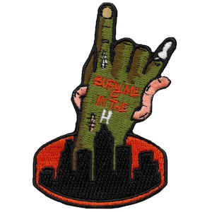 May include: Green zombie hand with a rock and roll hand gesture, emerging from a black cityscape with the text "BURY ME IN THE H" on the palm. The hand is surrounded by a red circle.