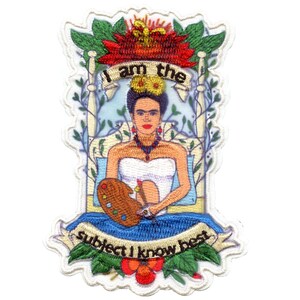 May include: Embroidered patch featuring Frida Kahlo in a white dress, holding a paintbrush and palette. The text "I am the subject I know best" is written on a blue ribbon.