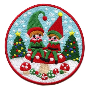 May include: Two elves wearing green and red hats sit on a red and white mushroom in a snowy winter scene. The elves are smiling and holding hands. There are two Christmas trees in the background, decorated with colorful lights and ornaments.