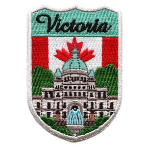 Op de afbeelding: Gestikt embleem met een wit schild op een groene achtergrond. Het schild heeft een rood-witte Canadese vlag met een rood esdoornblad in het midden. Het schild heeft ook een grijs gebouw met een groen dak en een fontein ervoor. De tekst "Victoria" is in het zwart bovenaan het schild geborduurd.
