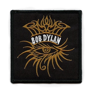 Peut inclure: Patch brodé noir avec un motif d'œil et de couronne dorés. Le texte "BOB DYLAN" est en lettres blanches.