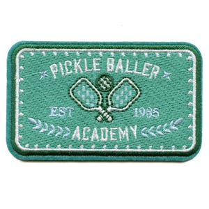 Może przedstawiać: Zielony haftowany znaczek z białymi ściegami. Zawiera rakietę do pickleballa i piłkę z tekstem "Pickle Baller Academy Est. 1985".