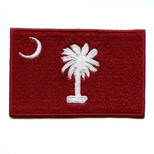 Puede incluir: Un parche bordado rojo con una luna creciente blanca y una palmera blanca. El parche es una representación de la bandera del estado de Carolina del Sur.