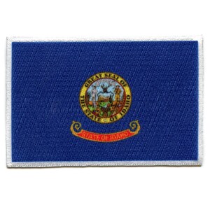 Op de afbeelding: Een blauwe patch met een witte rand met het staatszegel van Idaho. Het zegel toont een bergketen, een zon en een banner met de woorden "The State of Idaho".