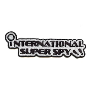 Puede incluir: Parche bordado en blanco y negro con la frase "INTERNATIONAL SUPER SPY". El parche tiene un borde blanco y un icono de lupa al principio. El texto está escrito en una fuente estilizada.