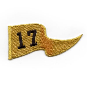 May include: Gold embroidered patch with the number 17 in black. The patch is shaped like a flag with a pointed end.