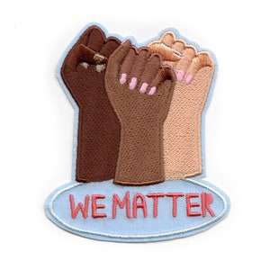 May include: Embroidered patch featuring three raised fists in varying skin tones. The fists are brown, tan, and dark brown, with pink nail polish. The patch has a light blue border and an oval base with the words "WE MATTER" in red.