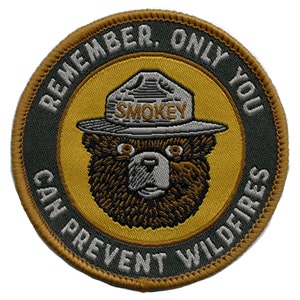 May include: A round, brown and green embroidered patch with the words "Remember, Only You Can Prevent Wildfires" surrounding a cartoon image of Smokey Bear wearing a ranger hat with the word "Smokey" on it.