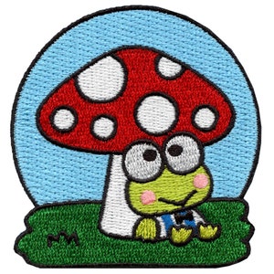 May include: An embroidered patch featuring a cartoon frog sitting under a red and white mushroom with white spots. The frog is wearing a blue and white striped shirt and a bow tie. The mushroom and frog are sitting on a green patch of grass.