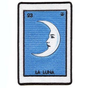 May include: A blue and white embroidered patch with the image of a crescent moon with a face. The text "LA LUNA" is below the moon and the number "23" is in the top left corner.
