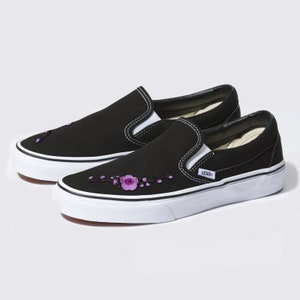 Könnte beinhalten: Schwarze Vans Slip-Ons mit weißer Sohle und lila Blumen Stickerei an der Seite. Die Schuhe haben ein Vans Logo an der Seite.