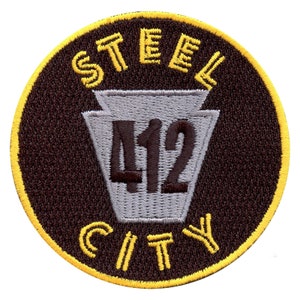 Puede incluir: Parche circular con las palabras "STEEL CITY" en amarillo, arqueadas en la parte superior e inferior. El número "412" está centrado dentro de una forma de clave de bóveda gris. El fondo es marrón oscuro con un borde amarillo.