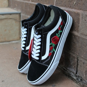 May include: Black and white Vans sneakers with red rose embroidery on the side. The shoes have white laces and a white stripe on the side.