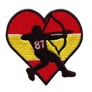Puede incluir: Parche en forma de corazón rojo, amarillo y blanco con una silueta negra de un arquero sosteniendo un arco y una flecha. El arquero lleva una camiseta con el número 87.