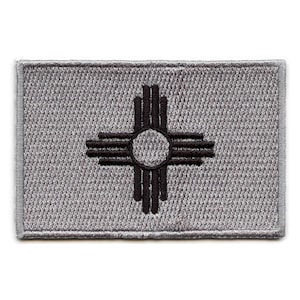 Op de afbeelding: Een grijze geborduurde patch met het staatssymbool van New Mexico, een zwart Zia-symbool, op een grijze achtergrond.