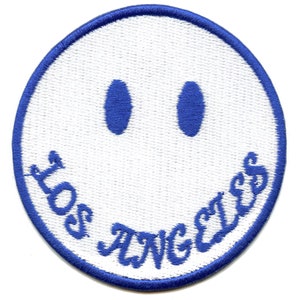 Op de afbeelding: Een witte patch met een blauwe rand en een lachend gezichtsontwerp. De ogen zijn blauwe ovalen en de tekst "LOS ANGELES" is in blauwe letters rond de glimlach geschreven.