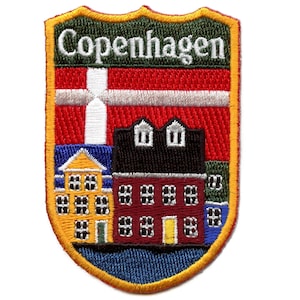 Pode incluir: Etiqueta bordada com um design em forma de escudo. A etiqueta apresenta uma bandeira dinamarquesa vermelha e branca, um edifício amarelo com janelas vermelhas e um edifício preto com janelas brancas. O texto "Copenhaga" está bordado em verde na parte superior da etiqueta.