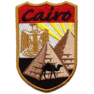 May include: Embroidered patch with the word "Cairo" in black lettering on a red background. The patch features a white background with a gold Egyptian eagle, a yellow sun, and two brown pyramids with a black camel in front of them.