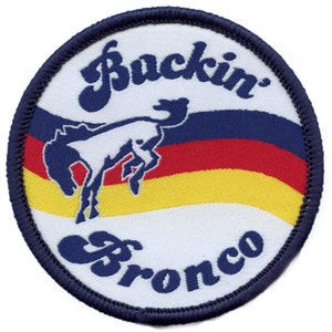 May include: A white circular patch with a blue border. The patch features a blue and white stylized horse with a red and yellow striped background. The text "Buckin' Bronco" is written in blue cursive.