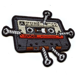 Könnte beinhalten: Gestickter Aufnäher mit einem grauen Kassettenband-Design mit dem Text "A June 27" und "Mixtape" in Weiß und Orange. Der Aufnäher ist mit stilisierten Schrauben verziert, was ihm eine einzigartige, Vintage-Ästhetik verleiht.