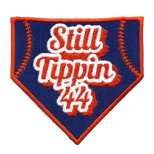 Puede incluir: Un parche en forma de base de béisbol azul y naranja con el texto "Still Tippin' 44" en letras blancas y naranjas.