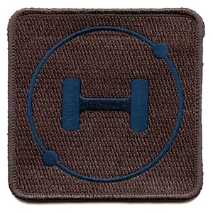 May include: A square, brown embroidered patch featuring a blue dumbbell inside a blue circle. The patch has a textured appearance and a brown border. The design suggests a fitness or gym theme.