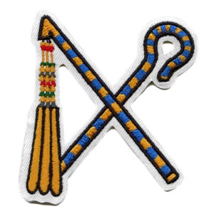 May include: A white embroidered patch with a crossed crook and flail, symbols of ancient Egyptian royalty. The crook is blue and gold, while the flail is gold with a red, green, and black tassel.