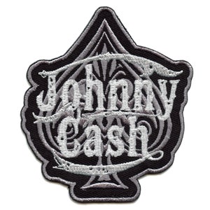Puede incluir: Un parche bordado en blanco y negro con el nombre "Johnny Cash" en una fuente estilizada. El parche tiene forma de pica y tiene un diseño decorativo alrededor de los bordes.