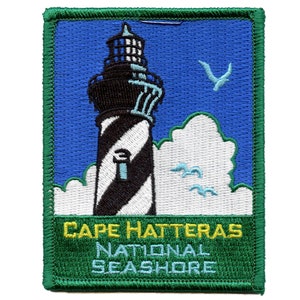 May include: A green embroidered patch with a white and black striped lighthouse, white clouds, and blue sky. The text "CAPE HATTERAS NATIONAL SEASHORE" is in yellow letters.