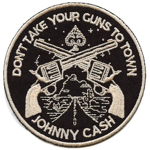 Puede incluir: Parche bordado en blanco y negro con el texto "Don't Take Your Guns To Town" y "Johnny Cash" alrededor de un diseño de dos pistolas cruzadas y una pica.