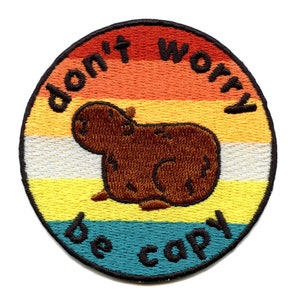 Puede incluir: Un parche redondo bordado con un fondo de arcoíris y una capibara marrón. El texto "don't worry be capy" está escrito en un contorno negro alrededor del borde del parche.