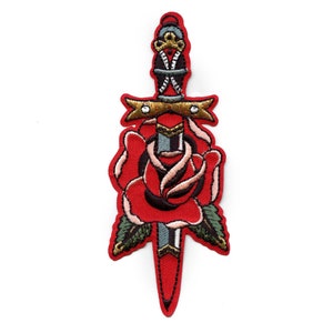 May include: A red rose with a silver dagger piercing through it. The dagger has a gold handle and a black cross on the blade. The rose has green leaves and a pink centre.