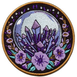 Op de afbeelding: Gestikt patch met een paarse kristallencluster in een glazen bol, omringd door paarse bloemen en bladeren op een donkerbruine achtergrond.