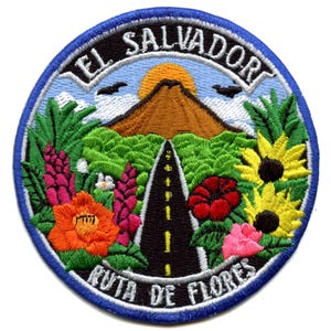 May include: Embroidered patch featuring a scenic landscape of a volcano with a road leading through a field of flowers. The text "EL SALVADOR" is written in a circle around the top of the patch, and the text "RUTA DE FLORES" is written in a circle around the bottom of the patch.