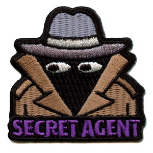 Könnte beinhalten: Gestickter Aufnäher mit einem Cartoon-Geheimagenten in einem Fedora und Trenchcoat. Das Gesicht des Agenten ist schwarz mit weißen Augen. Die Worte "SECRET AGENT" sind in Lila gestickt.