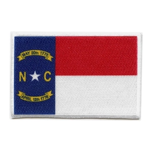 May include: A rectangular patch featuring the North Carolina state flag. The flag is blue with a white star in the centre and the letters "N.C." in white. The flag also has a yellow border with the words "MAY 20th 1775" and "APRIL 12th 1776". The flag is set against a white background.