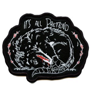 May include: Black embroidered patch featuring a stylized wolf curled up, with the text "IT'S ALL PRETEND" in white. The design includes red accents and a decorative border, suitable for jackets or bags.
