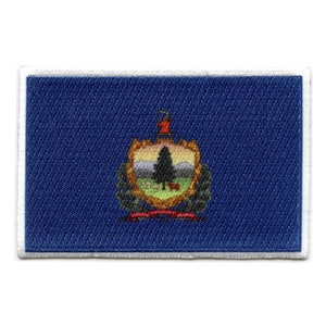 May include: A blue fabric patch with a white border, featuring the state seal of Vermont. The seal depicts a green pine tree on a green field with a red and white banner at the bottom.