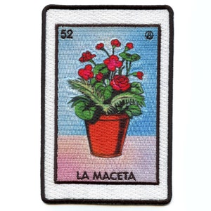 May include: A vintage-style embroidered patch featuring a red flower pot with green plants and red flowers. The patch has a white background with a blue sky and a pink and orange gradient at the bottom. The text "LA MACETA" is printed below the pot and the number "52" is printed in the top left corner.
