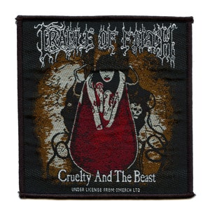 Op de afbeelding: Een zwarte en gouden geweven patch met de tekst "CRADLE OF FILTH" in een gotisch lettertype. De patch toont een vrouw in een rode jurk met een schedel in haar hand. De tekst "Cruelty And The Beast" is onder de afbeelding gedrukt. De tekst "UNDER LICENSE FROM OMERCH LTD" is onder de tekst gedrukt.