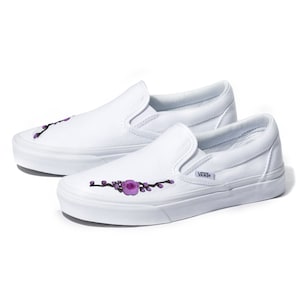 May include: White canvas slip-on Vans shoes with a purple floral embroidery design on the side. The shoes have a black Vans logo on the side.