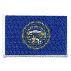 Op de afbeelding: Een blauwe geborduurde patch met een witte rand met het staatszegel van Nebraska. Het zegel toont een gouden ster met de woorden "Great Seal of the State of Nebraska" eromheen. Het zegel toont ook een afbeelding van een rivier, een brug en een boom.