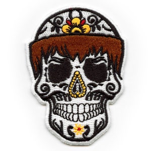 May include: Embroidered white skull with brown hair, black eyes, and a white flower with a yellow center on the chin. The skull is wearing a white headband with a yellow and red flower design.