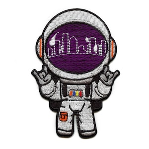 May include: Embroidered iron-on patch of a cartoon astronaut wearing a white spacesuit with a purple visor and a city skyline inside. The astronaut is giving the 'hang loose' hand gesture with both hands.
