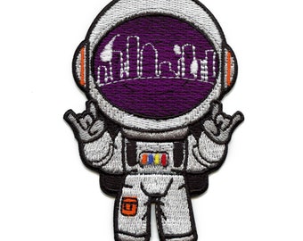 May include: Embroidered iron-on patch of a cartoon astronaut wearing a white spacesuit with a purple visor and a city skyline inside. The astronaut is giving the 'hang loose' hand gesture with both hands.