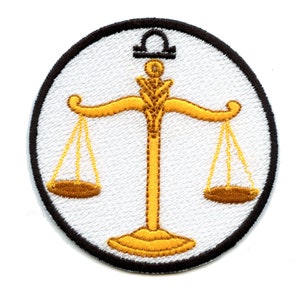 Op de afbeelding: Een witte geborduurde patch met een zwarte rand, met een gouden weegschaal symbool. De weegschalen zijn in balans en hebben een gouden basis.